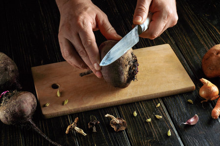 A person is peeling a beet using a knife on a wooden cutting board. Other vegetables like potatoes...