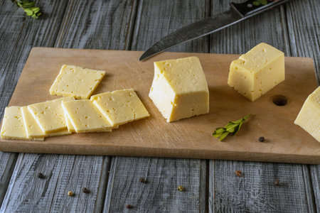 Cheese is cut into slices and placed on a wooden board. A knife is next to the cheese, and various...