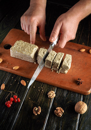 A person cuts a block of nougat into pieces on a wooden cutting board. Nuts and spices are placed...