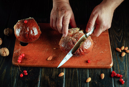 A person is cutting round bread on a wooden board with a knife. Nearby, there are walnuts and...