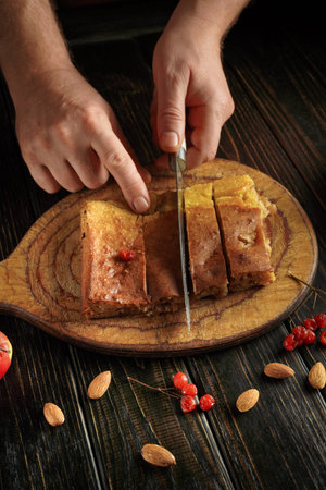 A person uses a knife to cut a dessert on a wooden board. Fresh fruit and nuts are placed around the...