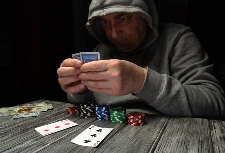 A man sits at a table holding playing cards while focused on the game. He has poker chips and cash...