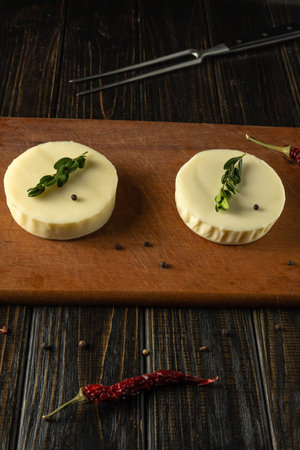 Two rounds of fresh cheese sit on a wooden cutting board. Each piece has a green herb and a black...