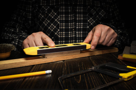 A man holds a yellow level tool against a piece of wood in a workshop. He appears focused as he measures the wood surface. A pencil and other tools are nearby on a wooden table.