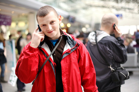 Young man in a red jacket calling on the phone at the airport. another caller man in the background.の写真素材