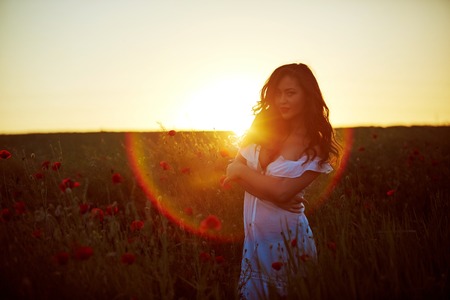 Beautiful woman in a poppy field with flowers on the sunset. Woman wearing in a white dress.の写真素材