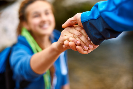 Helping hand - hiker woman getting help on hike smiling happy overcoming obstacle. Tourist backpackers walking in autumn forest. Young couple traveling.