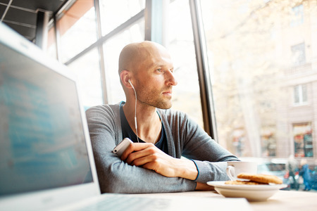 joyful smiling man sitting in a cafe near the open laptop at a table made of wood and holds a smart phone with headphones. in the background a bright window with bright daylightの写真素材