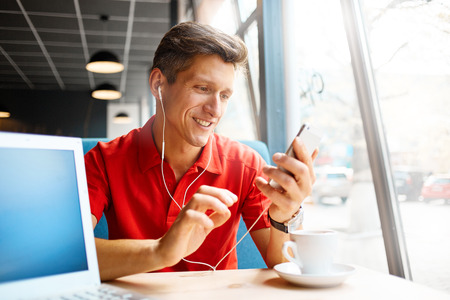 joyful smiling man sitting in a cafe near the open laptop at a table made of wood and holds a smart phone with headphones. in the background a bright window with bright daylightの写真素材