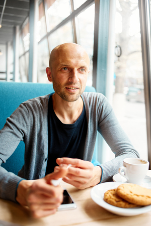 joyful smiling man sitting in a cafe near the open laptop at a table made of wood.  in the background a bright window with bright daylightの写真素材