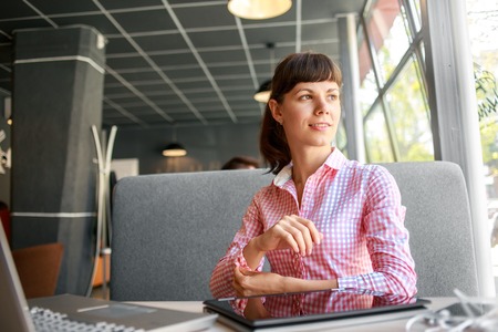 joyful smiling woman sitting in a cafe near the open laptop at a table made of wood and holds a smart phone with headphones. in the background a bright window with bright daylightの写真素材