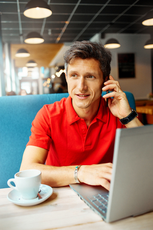 joyful smiling man sitting in a cafe near the open laptop at a table made of wood.  in the background a bright window with bright daylightの写真素材
