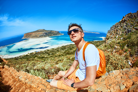 male traveler with backpack  sitting on the trail against sea and blue sky at early morning. Balos beach at backgroundの写真素材