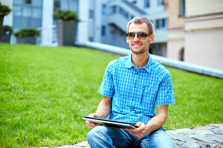 young man, wearing in blue t-shirt and sunglasses  with thin, elegant flexible laptop  on the green background in the park or business center or univercity area. learning or business conceptの写真素材