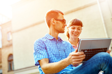 young man, wearing in blue t-shirt and sunglasses and woman with thin, elegant flexible laptop on the green background in the park or business center or university area.の写真素材