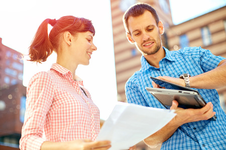 young man, wearing in blue t-shirt and sunglasses and woman with thin, elegant flexible laptop on the green background in the park or business center or university area.の写真素材