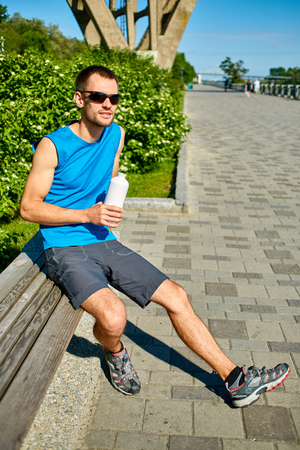 man runner athlete warming up and resting before jogging along a green bushes in the early morning. a man sitting on a bench with a bottle of water. man fitness sunset jogging workout wellness concept.の写真素材