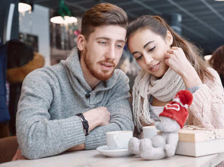 Two people, man and woman in cafe communicate, laughing and enjoying the time spending with each other. Couple in love on a date. A woman receives a gift from a man. Presented with a teddy bear in a red cap. Love story and Valentines Day conceptの写真素材