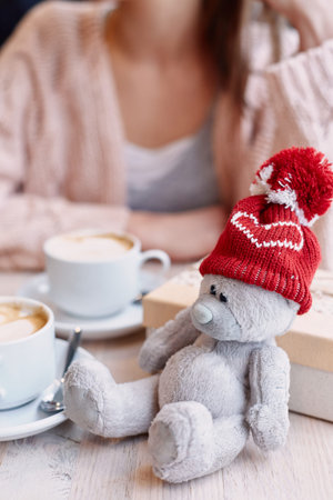 Teddy bear in red cap given on Valentines Day. smiling woman in cafe on background. Box with a gift and Two cups of coffee on table. bear in focusの写真素材