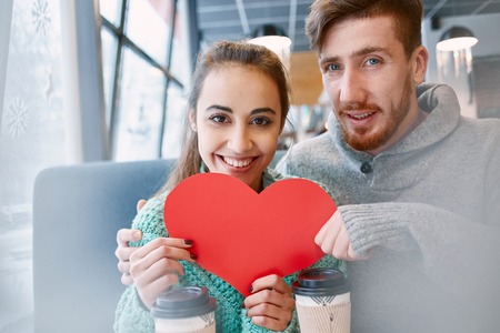 man and woman smiling and holding a large paper heart. Two people, man and woman in cafe communicate, laughing and enjoying the time spending with each other. Couple in love on a date. Love story and Valentines Day conceptの写真素材