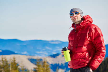 hiker in red down jacket with big plastic cup resting near the tent in the Carpathians mountains at...