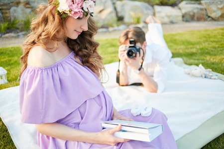 Pregnant woman and her husband sits on the grass in park at evening. Family enjoying pregnancy. man makes photo of woman by the camera. The concept of Mothers Day and Womens Day.の写真素材