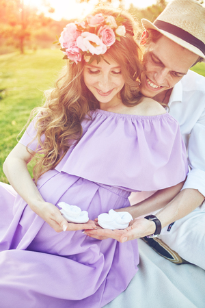 Pregnant woman and her husband sits on the grass in park at evening. Family enjoying pregnancy. man embracing a woman from behind and gives her a little baby slippers. The concept of Mothers Day and Womens Day.の写真素材