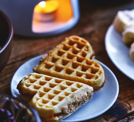 Two wafer cakes in the shape of heart lie on a small plate on a served wooden table. Next to a plate of sliced bananas, a bowl of cottage cheese and a fondue with chocolateの写真素材