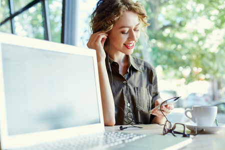 joyful smiling woman sitting in a cafe near the open laptop at a table made of wood . in the background a bright window with bright daylight. Woman listening to music from her phone in headphonesの写真素材