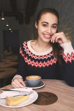 young woman at cafe drinking coffee and smiling. woman sits alone at the wooden table. Woman dressed in a knitted sweaterの写真素材