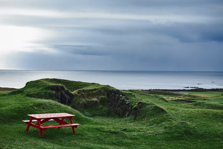Red old homemade wooden bench and table on the green lawn near the ancient ruins of a house on the ocean background in Icelandの写真素材