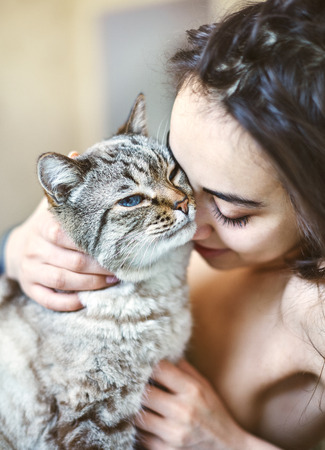 Happy beautiful young woman in the morning in bed with a cat. A woman without clothes and covered with a blanket. Morning photograph with a natural light from the window at home. Good mood and a pleasant start to the dayの写真素材