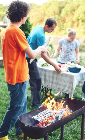 flame grilled and wood burning in fire and people on BBQ party. family barbecue party in outdoor or home garden. A man blows coals in a fire with a sheet of cardboard.の写真素材