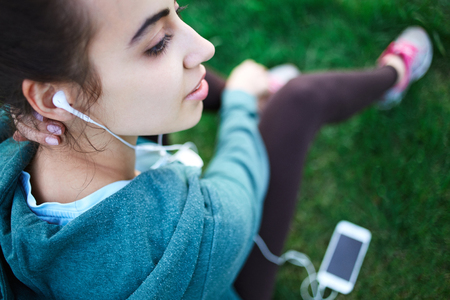 Portrait of young and sporty woman in sportswear sitting with the smartphone and headphones on the grass outside at the park on green field on cloudy day, Dnipro, Ukraine. She is sits on the green meadow between trees and listen musicの写真素材