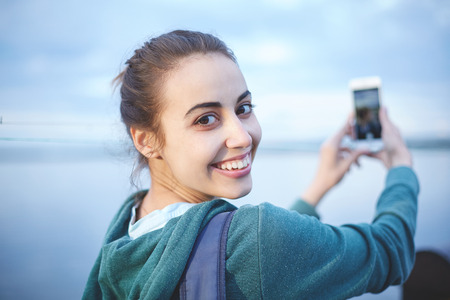 Portrait of beautiful happy girl in sportswear with small urban backpack on the shore of a lake or sea. woman makes selfie photo or video call.の写真素材