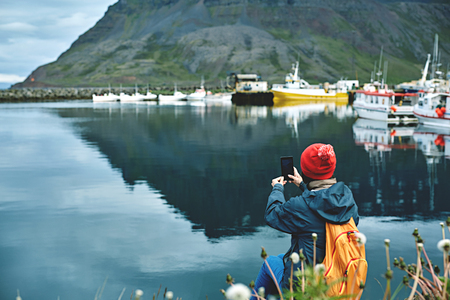 photo of girl in warm clothing with backpack on pier with background of mountains of West Fjords, Iceland. woman takes a photo on smartphoneの写真素材