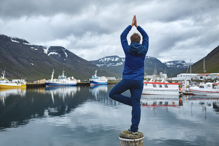 Back view of girl in warm clothing in yoga pose on pier with background of port and mountains of West Fjords, Bolungarvik, Iceland.の写真素材