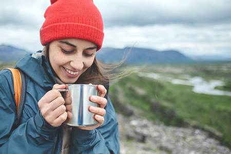 Girl in warm clothing and red hat hold a iron cup and drink a hot tea on background of mountains of Iceland.の写真素材