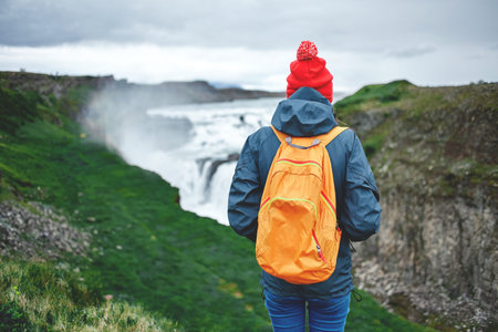 Girl in warm clothing, in red knitted hat and small orange backpack stands on the cliff on background of Gullfoss waterfall in Iceland. Travel to the Icelandの写真素材