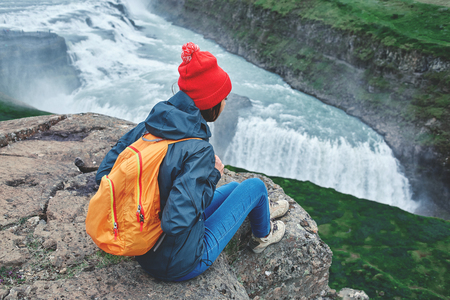 Girl in warm clothing, in red knitted hat and small orange backpack sits on the cliff on background of Gullfoss waterfall in Iceland. View from aboveの写真素材