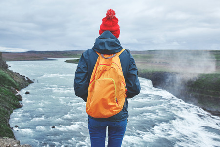 Girl in warm clothing, in red knitted hat and small orange backpack stands on the cliff on background of Gullfoss waterfall in Iceland. Travel to the Icelandの写真素材