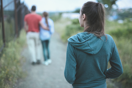back view of romantic couple on the trail at the summer at sunset. A single lonely woman is walking behind and looking at a coupleの写真素材