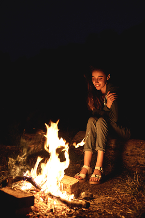 Young woman in geans and hoodie in the mountains. Young woman with fire on mountain at sunset. Girl standing near a fireplace, back viewの写真素材