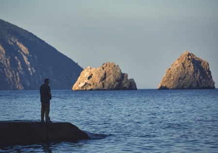 Side view of male fisherman standing on a pier with a fishing rod at sunsetの写真素材