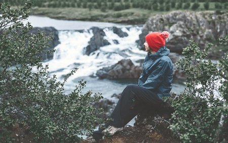 a woman tourist in waterproof clothes and red hat sits on the Bank of the mountain river with small waterfall. Glanni waterfall, Icelandの写真素材