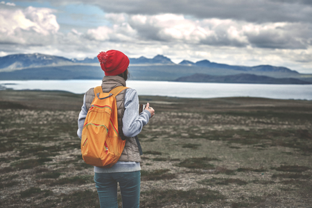 Girl dressed in a sweatshirt, a vest and a red knitted hat hold a iron cup and drink a hot tea on background of mountains and lake. Valley and national park Thingvellir, Icelandの写真素材