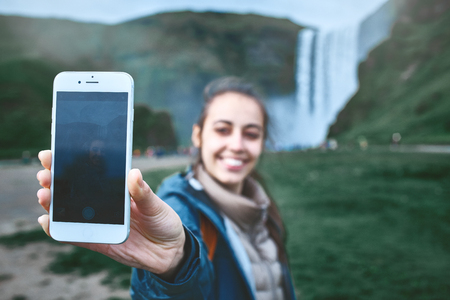 smiling woman in warm clothes making selfie on a Skogafoss waterfall background, Iceland. Focus on the phoneの写真素材
