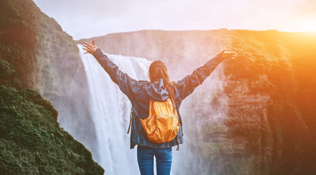 Girl in waterproof clothing stands on the cliff on background of Skogafoss waterfall in Iceland. back view, woman holds hands upの写真素材