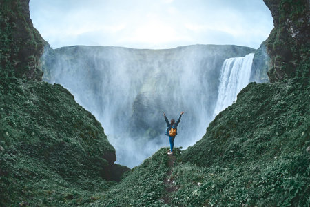 Girl in waterproof clothing stands on the cliff on background of Skogafoss waterfall in Iceland. back view, woman holds hands upの写真素材