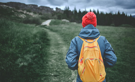 back view of young woman hiker hiking in beautiful mountains. female hiker in waterproof clothes, red hat and with small orange packpack on the green pines backgroundの写真素材